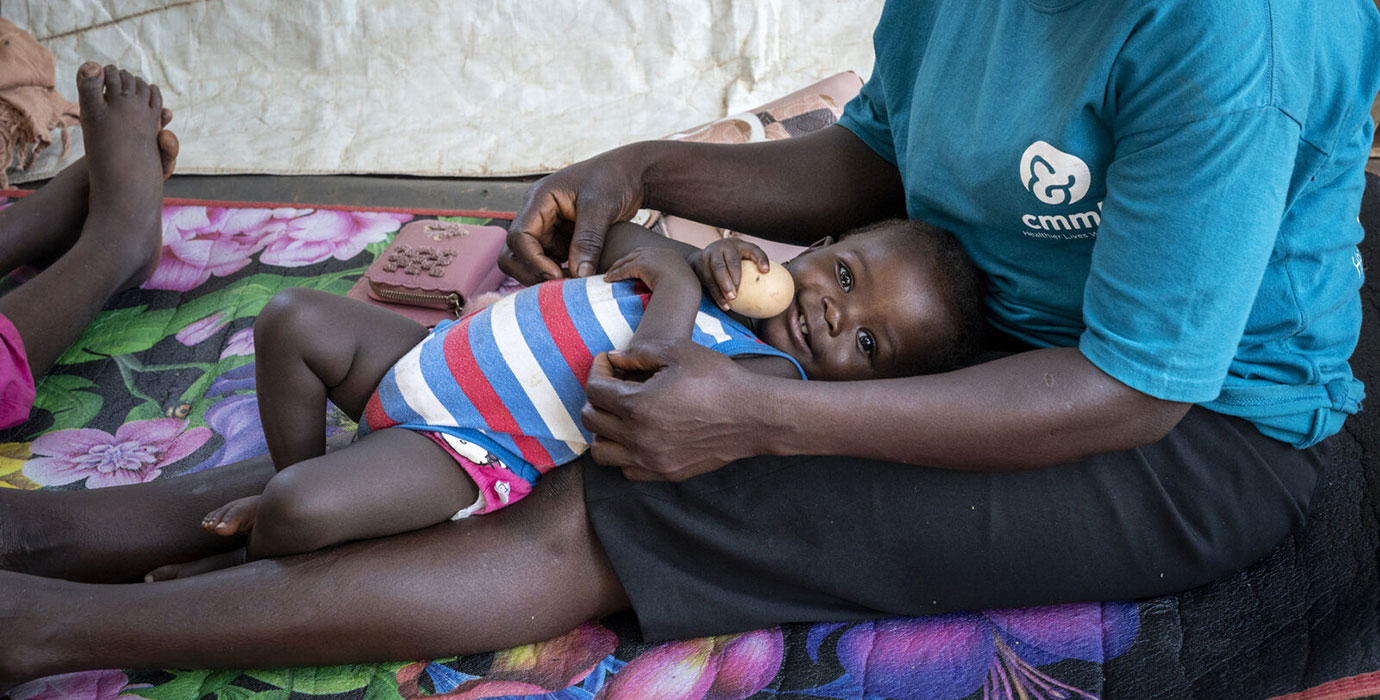 A boy smiles from his mothers lap, holding an egg.
